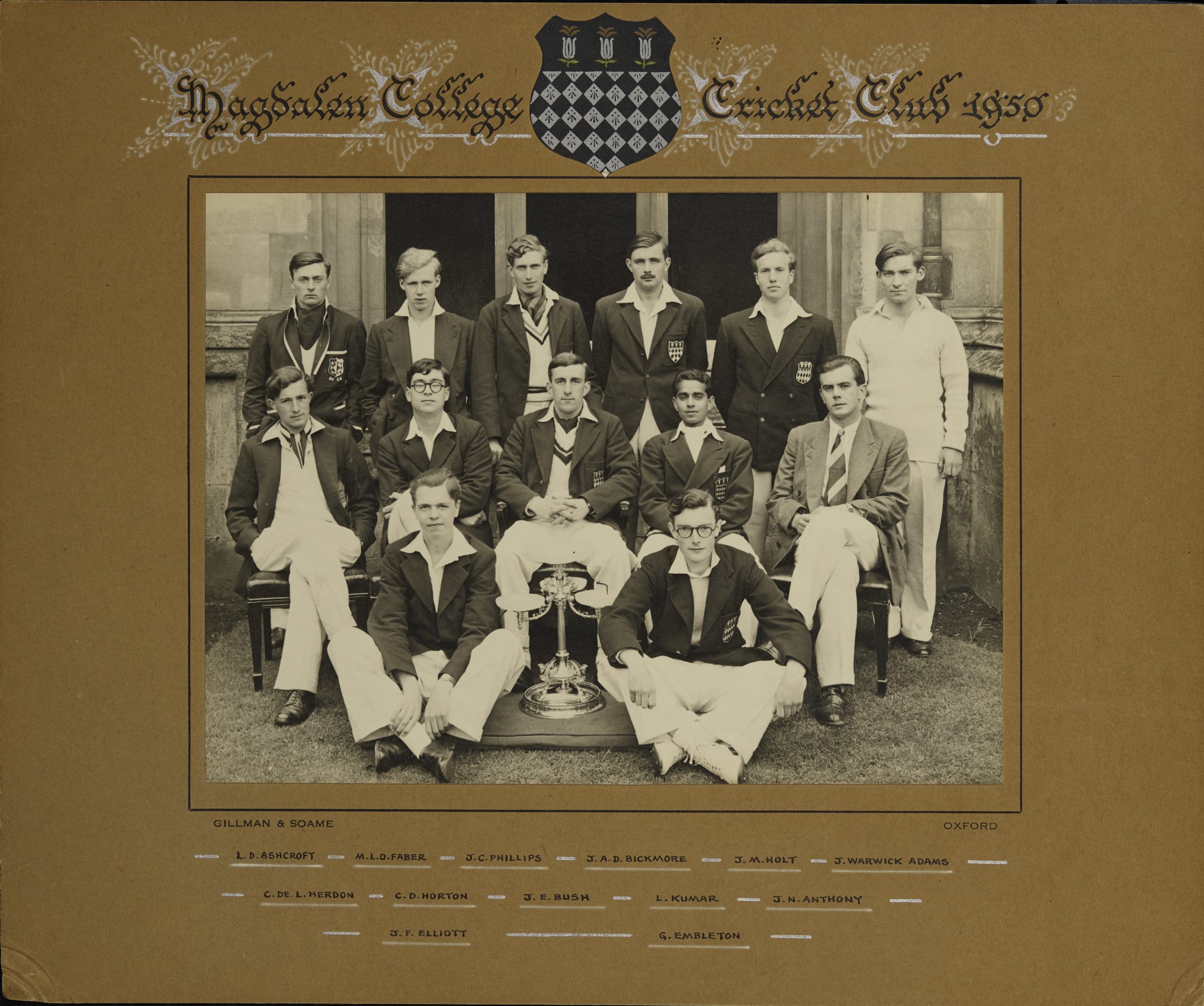 Black and white photograph of the cricket team, posed in the cloisters. 6 men are standing, 5 (including Lovraj Kumar) are sitting in chairs in front of them, and finally 2 men are sitting cross-legged at the front. Between the two men at the front is some kind of metal object- possibly a trophy. In the frame of the photo, the names of the men are handwritten in all caps, according to their position on the photo, thus: Back row: L. D. Ashcroft, M. L. D. Faber, J. A. D. Bickmore, J. M. Holt, and J. Warwick Adams. Middle row: C. de. L. Herdon, C. D. Horton, J. E. Bush, L. Kumar, J. N, Anthony. Front row: J. F. Elliott and G. Embleton.