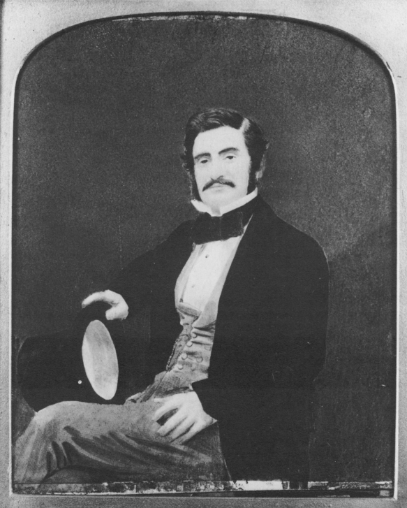 Black and white portrait of Hormuzd Rassam in Western formal wear. He is sitting and looks directly at the camera. He is wearing a bow tie and suit, and holding a top hat in his left hand